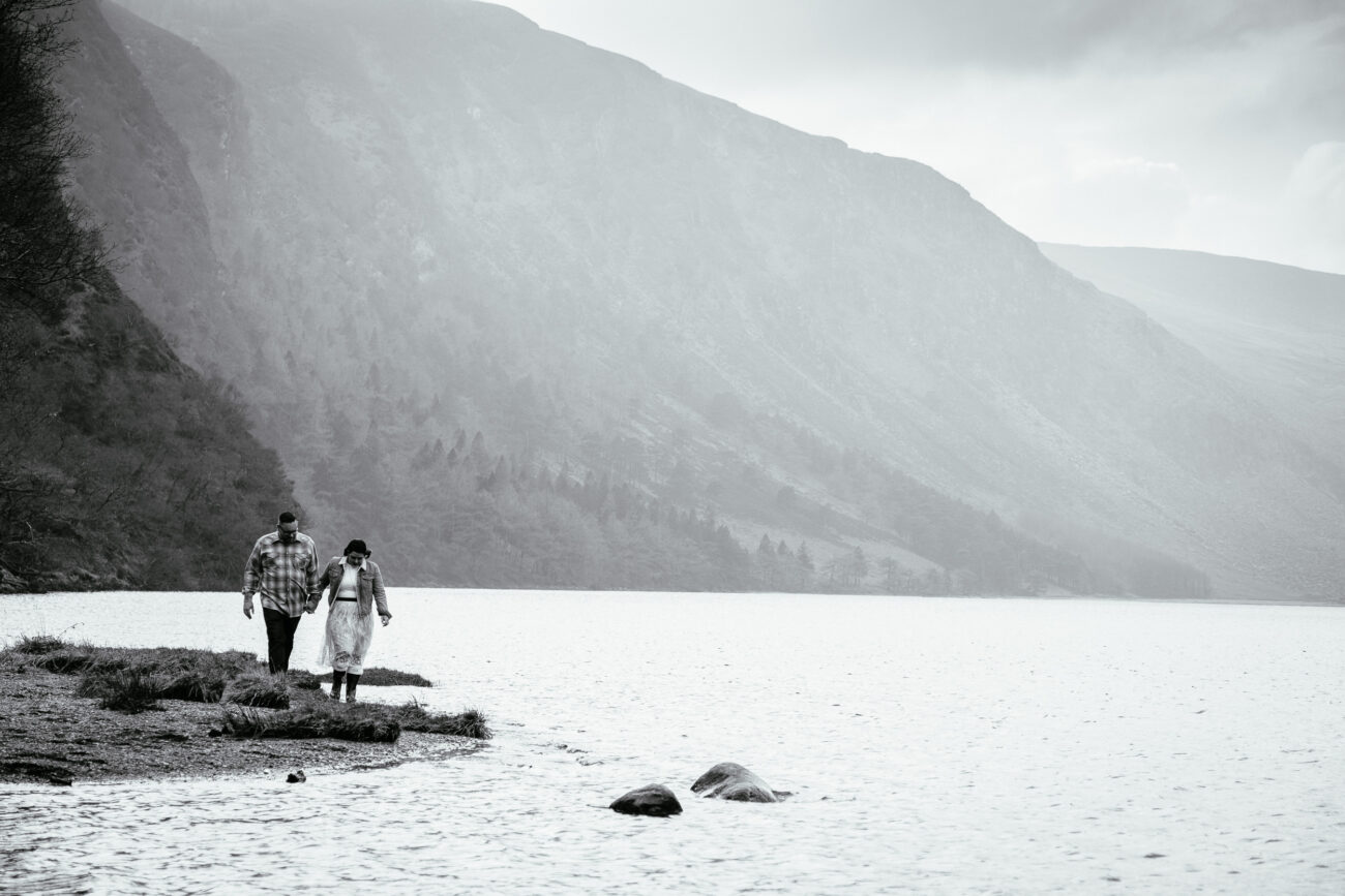 Two individuals walk on a grassy path beside a large body of water, with a mountain in the background and fog overhead.