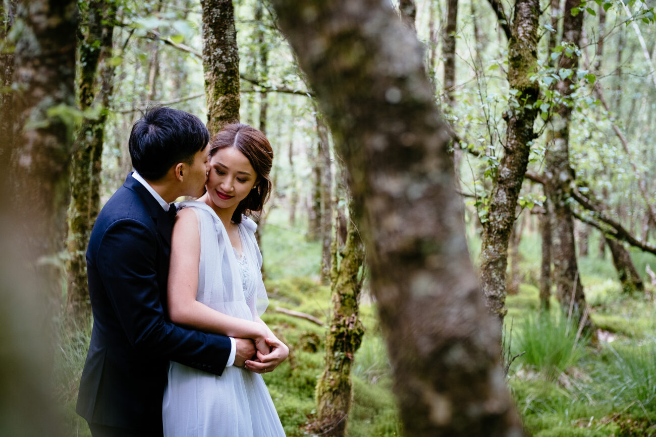 A couple wearing wedding attire embraces in a wooded area. The groom is dressed in a black suit, and the bride is in a white gown. Trees and greenery surround them.