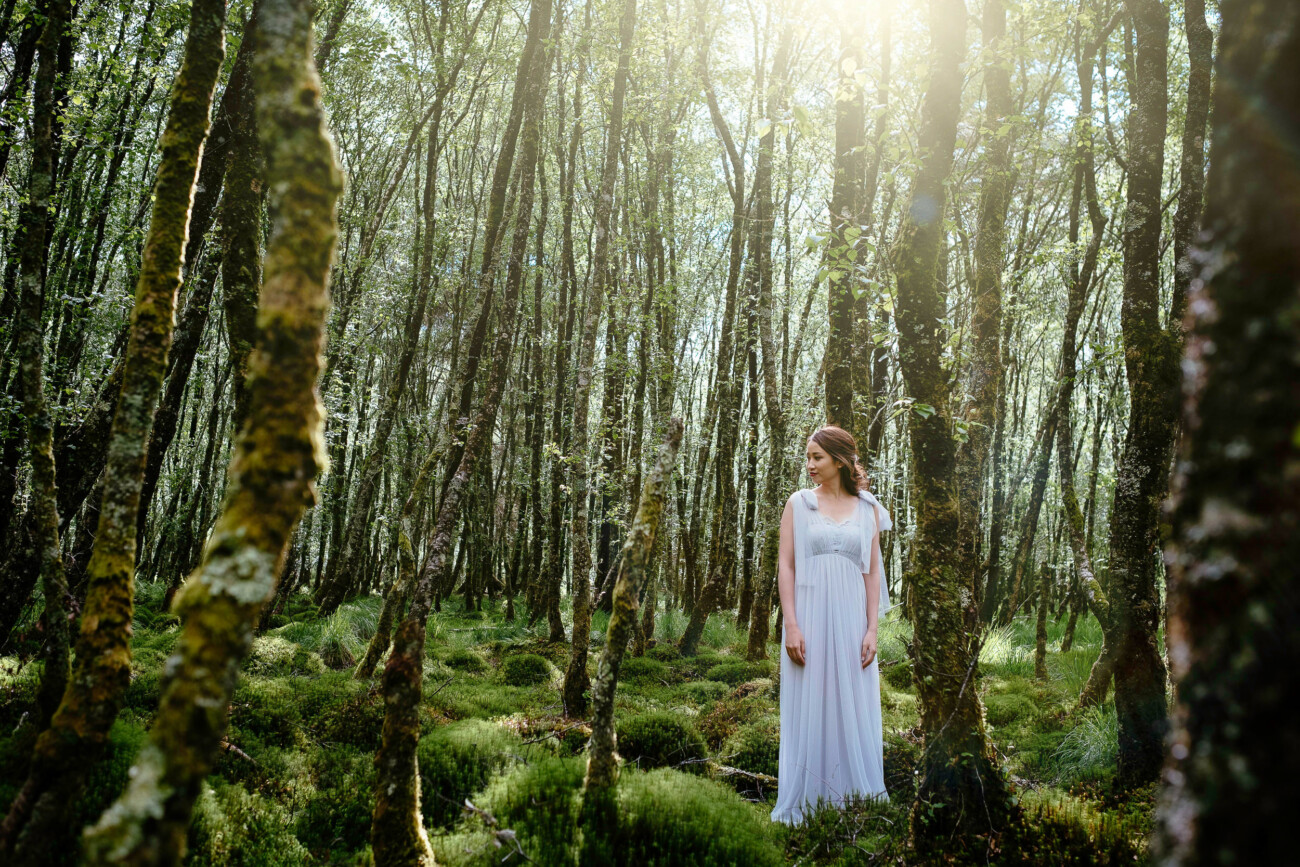 A woman in a white dress stands among mossy trees in a sunlit forest, capturing the magic of engagement photos in Ireland.