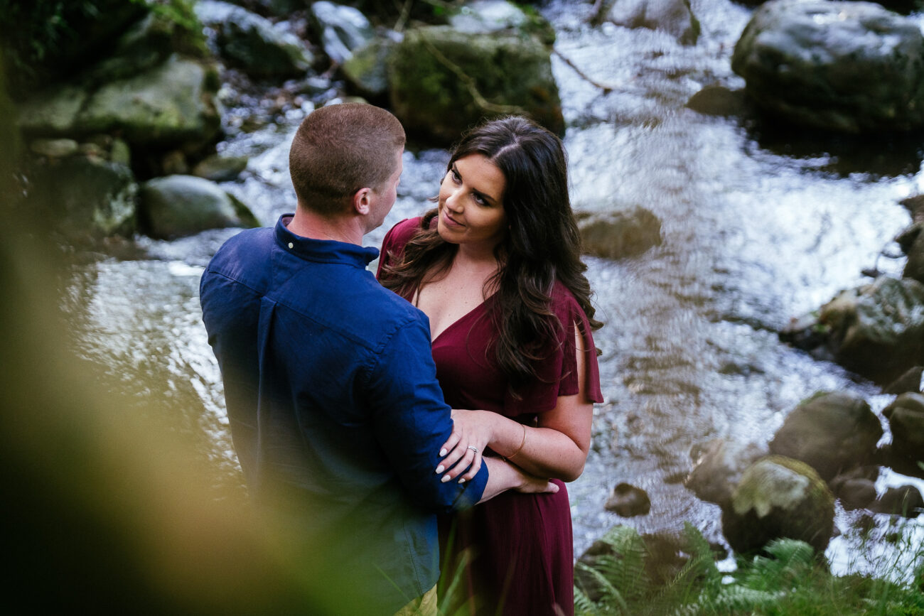 A man in a blue shirt and a woman in a red dress stand close together by a rocky stream, looking at each other.