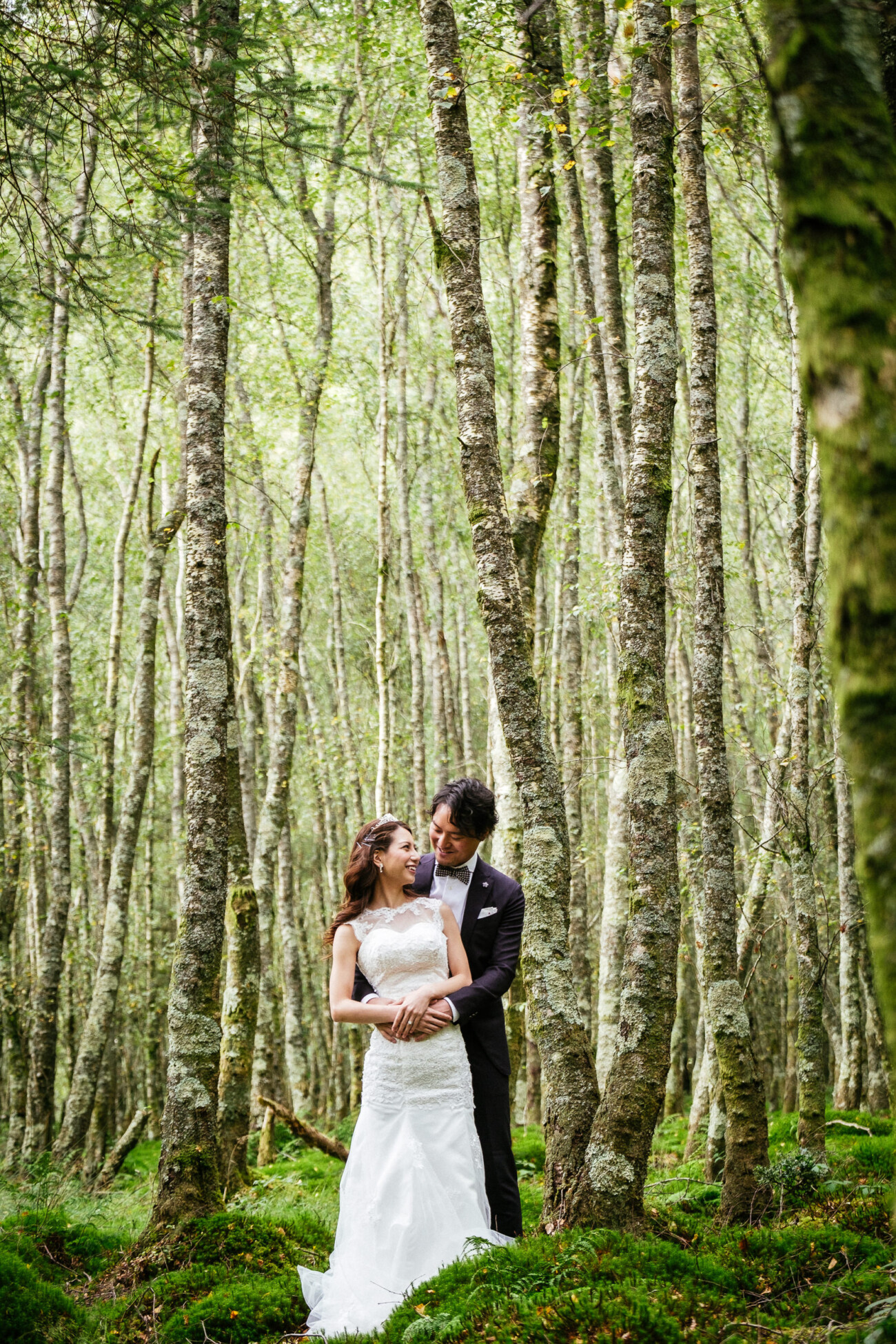 A bride and groom stand together in a forest, surrounded by tall, slender trees and green foliage—capturing the magic of Engagement Photos in Ireland.