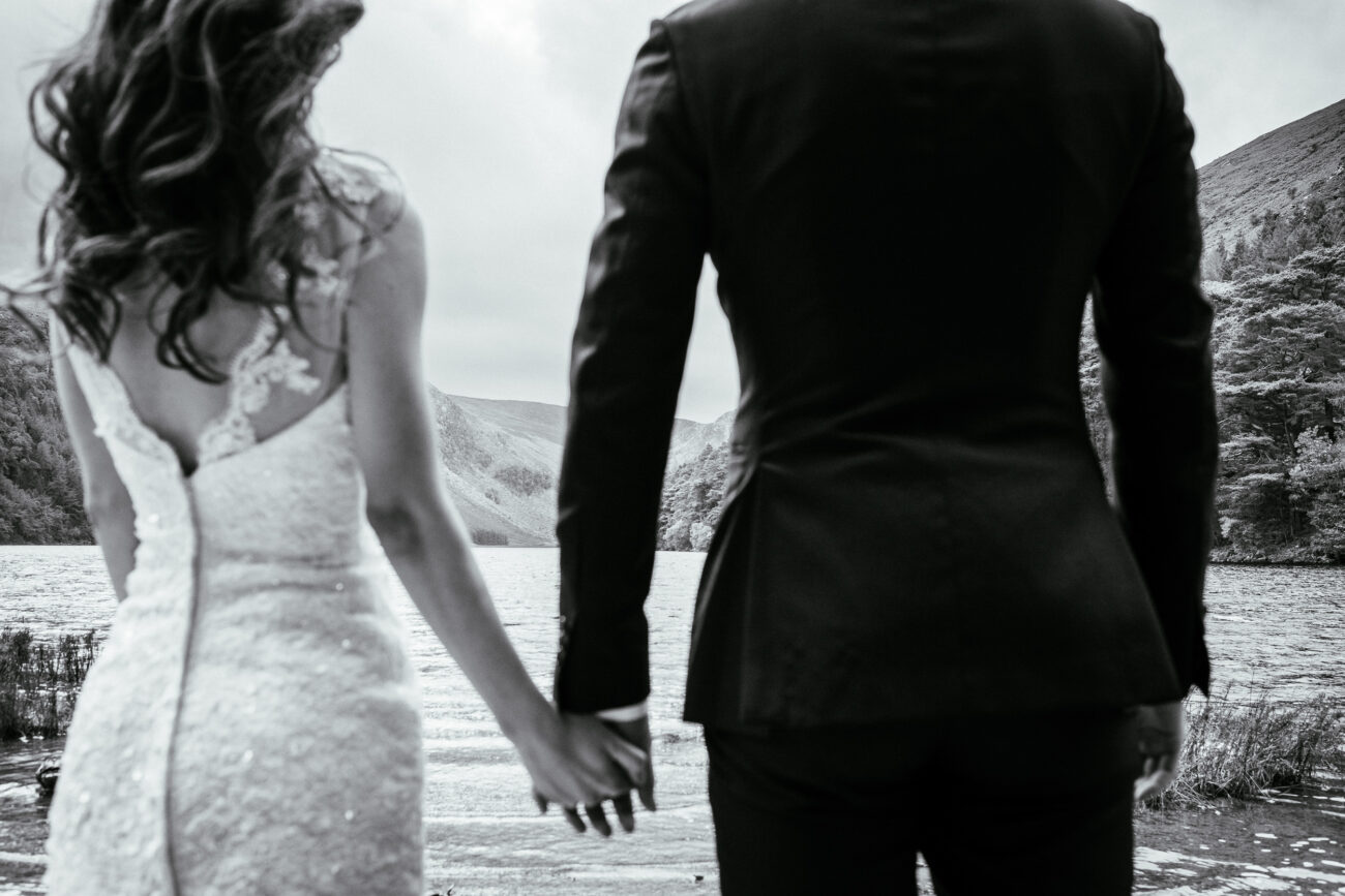 A couple in formal attire stand hand in hand facing a lake with mountains in the background.