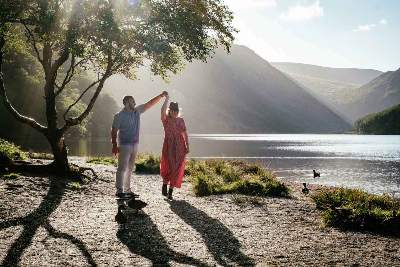 A man twirls a woman in a pink dress near a lake, surrounded by mountains and trees, with sunlight streaming through the branches—capturing the magic of engagement photos in Ireland.