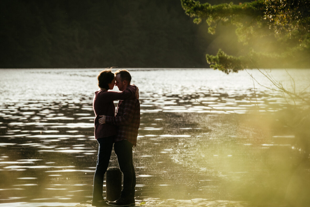 A couple embraces while standing near the edge of a calm lake, with sunlight reflecting on the water and trees in the background.