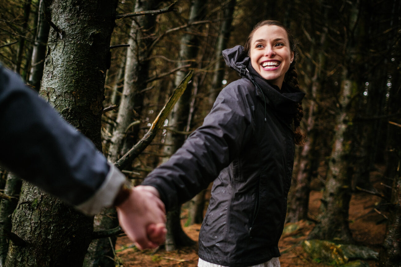 A person wearing a dark jacket smiles while holding hands with another person in a forest.