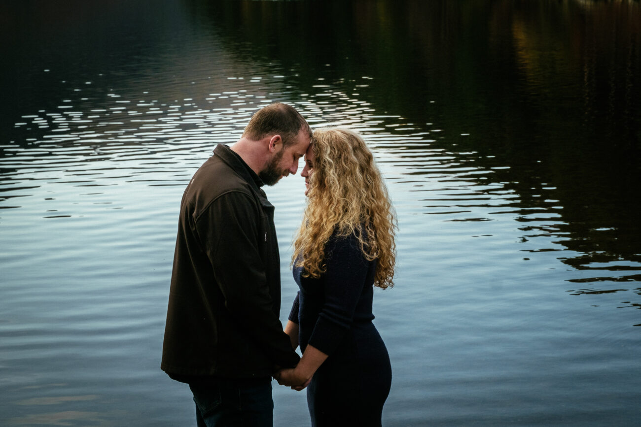 A man and woman stand close together, holding hands and touching foreheads by a calm body of water.