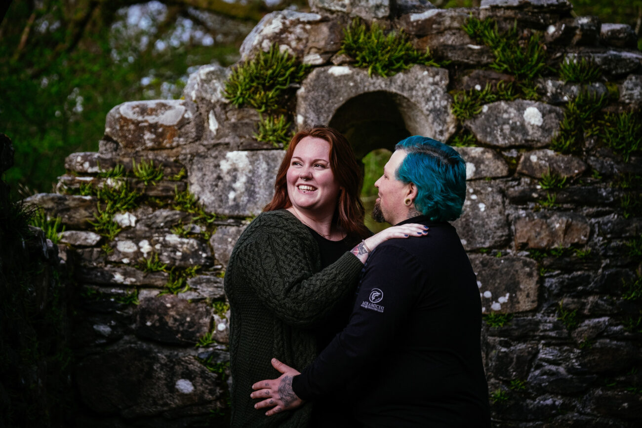 Two people, one with red hair and the other with blue hair, are standing close together in front of an aged, stone wall covered with patches of greenery.