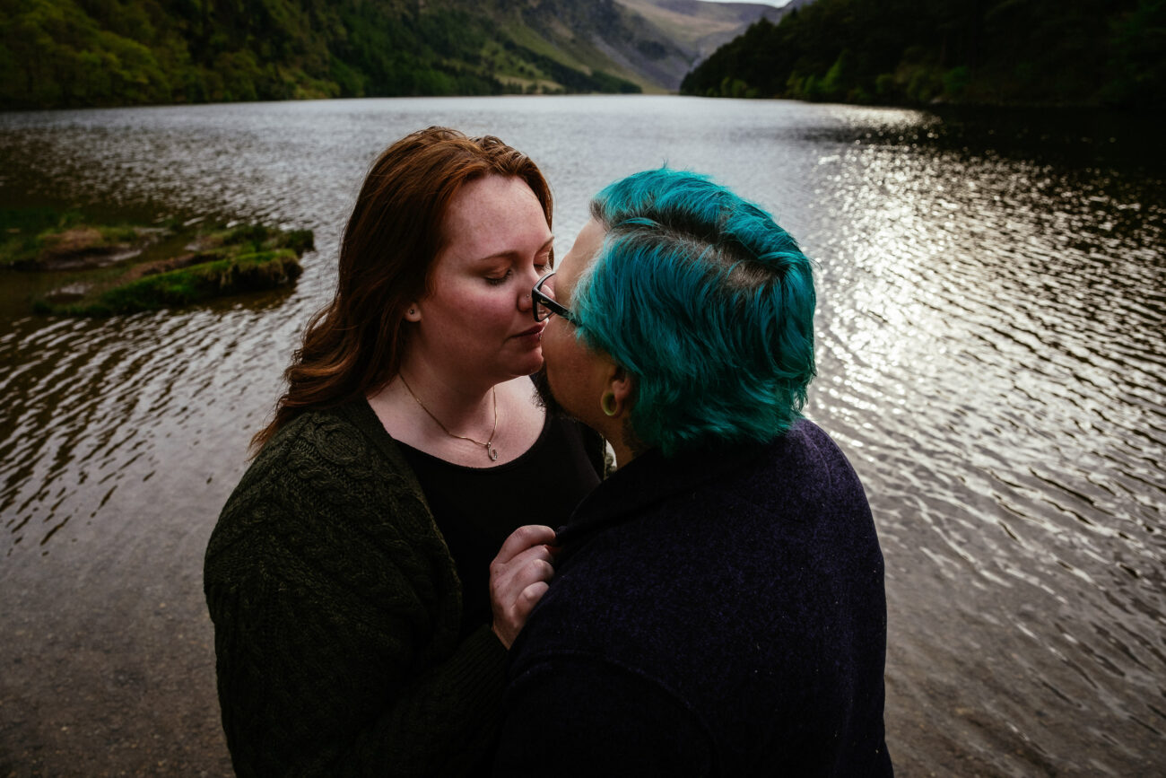Two people with close-cropped turquoise hair and long brown hair appear to be about to kiss while standing close to a lakeside, surrounded by a forested area with mountains in the background.