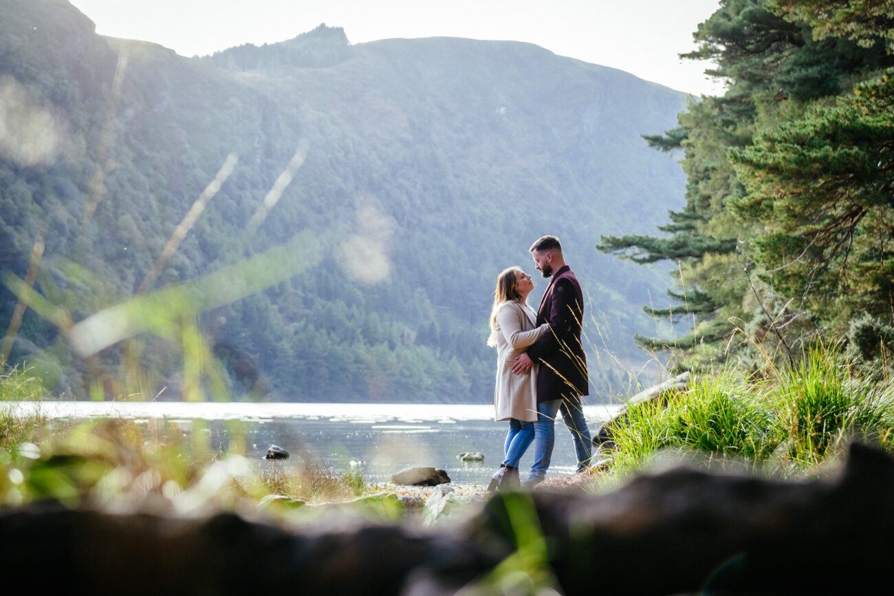 Two people stand close together outdoors near a lake with mountains in the background. They appear to be embracing, surrounded by greenery.