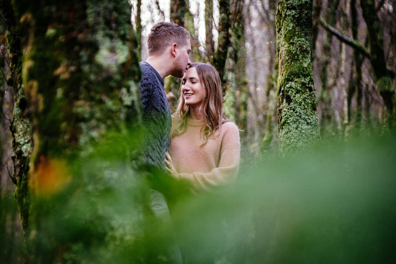 A couple stands in a forest; the man kisses the woman's forehead as they smile, surrounded by moss-covered trees and blurred green foliage in the foreground.