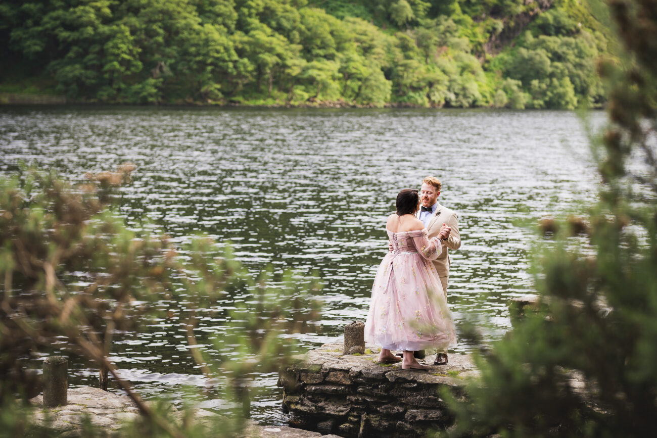 A couple dressed in formal attire stands on a stone platform by a serene lake, surrounded by lush green trees. The woman wears a floral dress, and the man is in a beige suit.