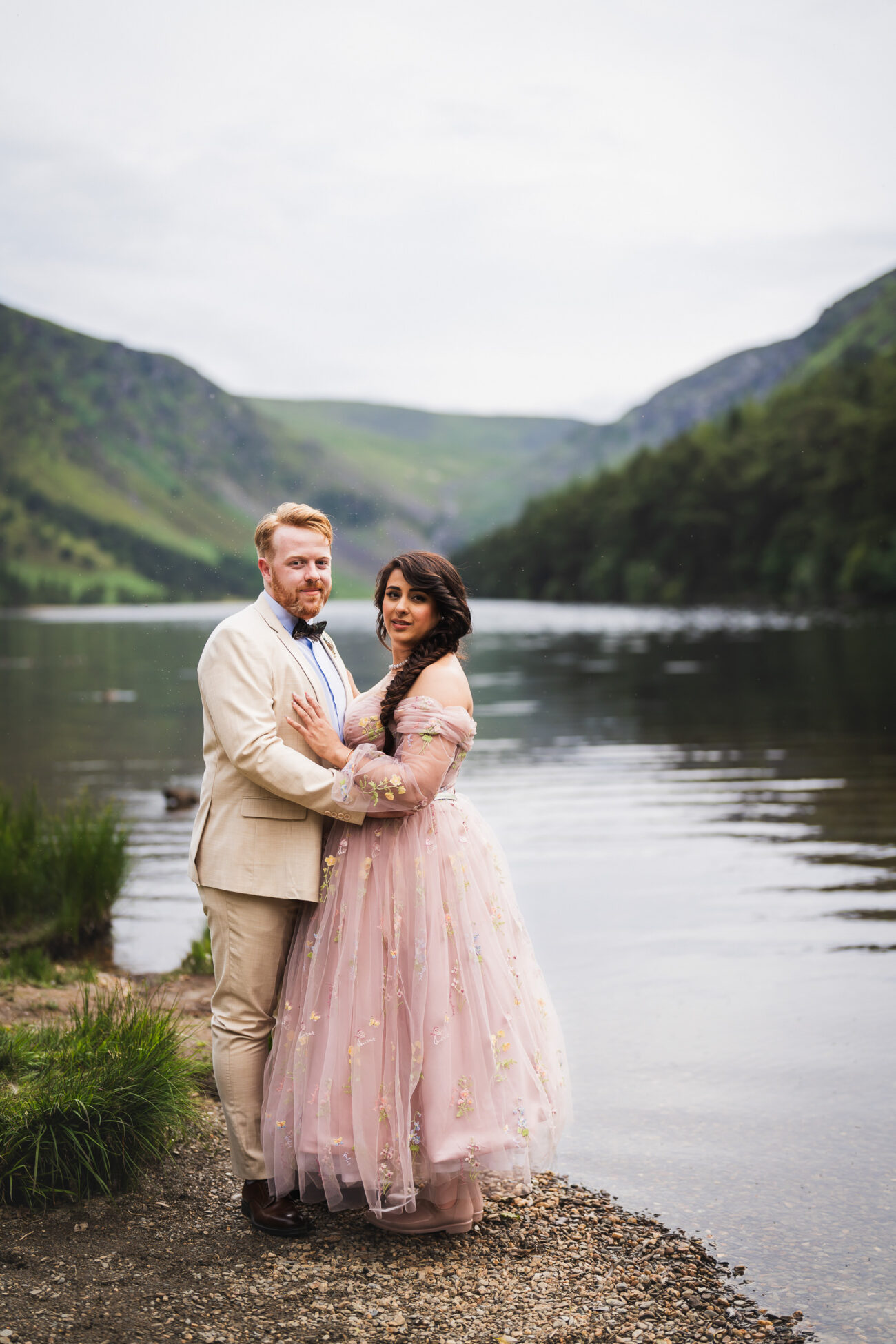 A couple stands together by a lake, surrounded by mountains and greenery. The woman wears a flowing pink dress, and the man wears a light-colored suit.