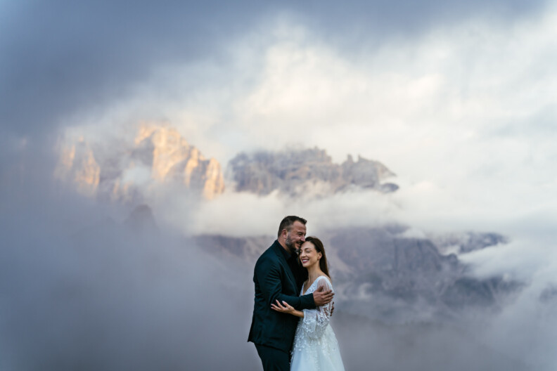 A couple in wedding attire embraces with misty mountains in the background.