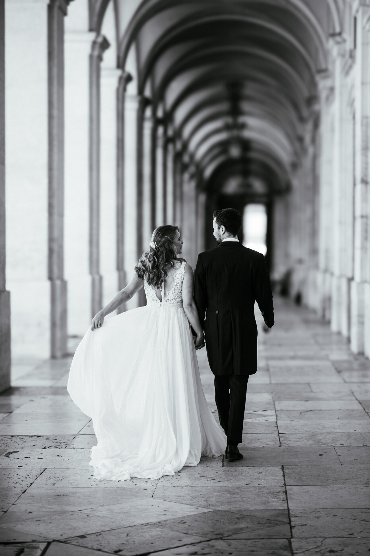A bride and groom walk hand in hand through an arched hallway, with the bride holding her dress. The photo is in black and white.