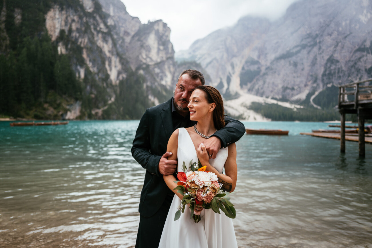 A couple stands by a mountain lake. The woman holds a bouquet of flowers, while the man embraces her from behind. Majestic mountains are visible in the background.