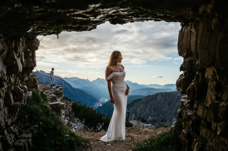 Woman in a white dress stands in a rocky cave opening with a mountainous landscape and cloudy sky in the background.