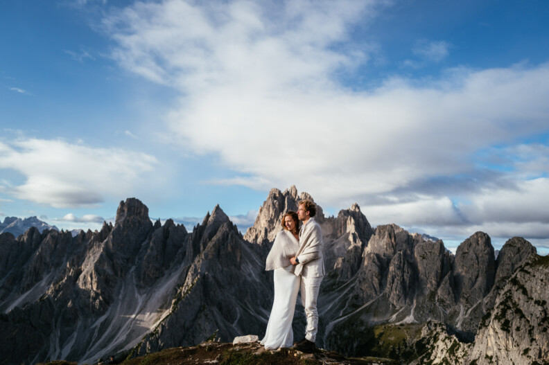 A couple dressed in white stands on a mountain with jagged peaks in the background under a blue sky with clouds.