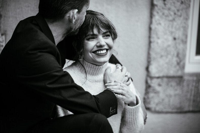 A couple sits closely, with the man whispering in the woman's ear. She is smiling broadly, wearing a light sweater. The background features a stone wall and window.