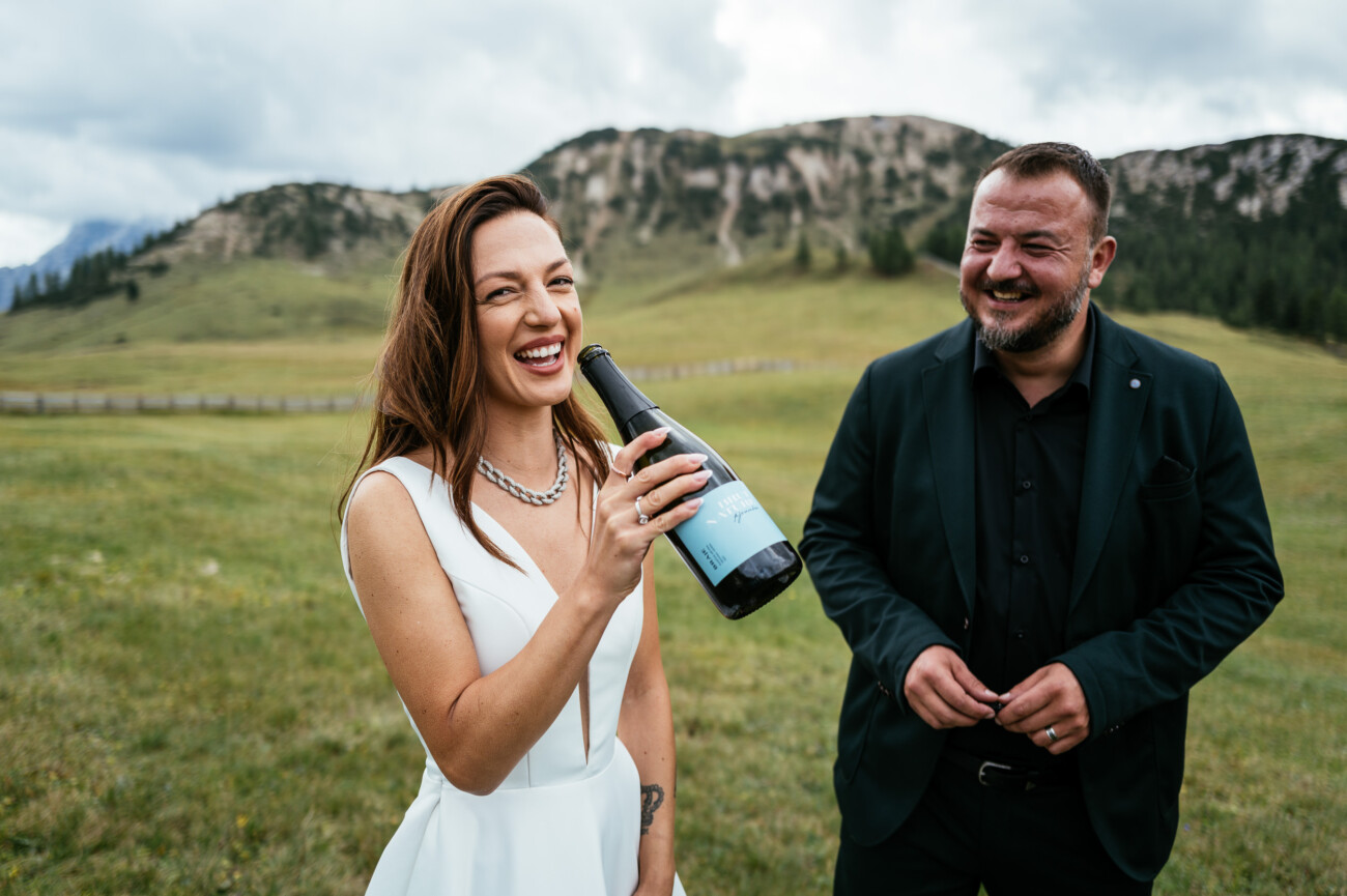 A woman in a white dress holds a wine bottle and laughs beside a smiling man in a dark suit. They are standing in a grassy field with hills and cloudy sky in the background.