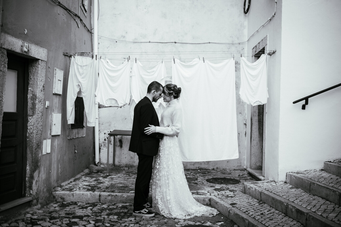 A couple in wedding attire embraces in front of a clothesline with white sheets in a narrow alley.