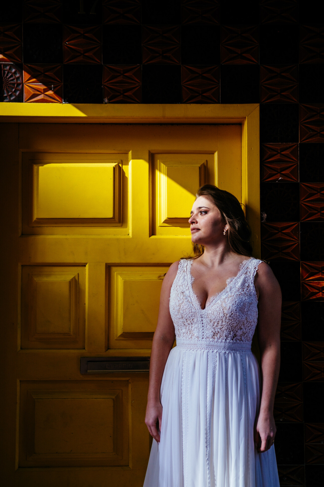 A woman in a white dress stands against a yellow door with patterned tiles, looking to the side.