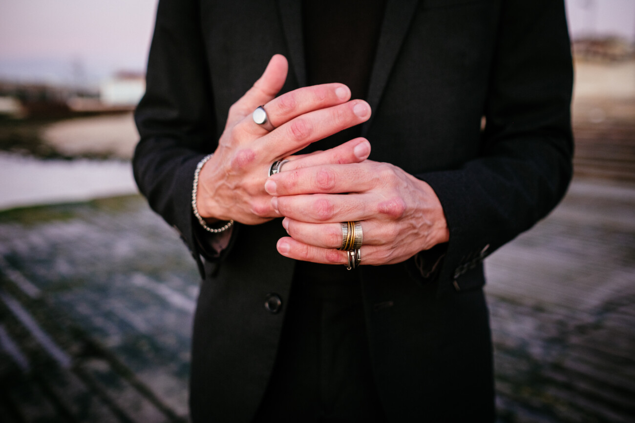 Person in a black suit showing their hands with multiple rings and bracelets, standing outdoors on a wooden surface.
