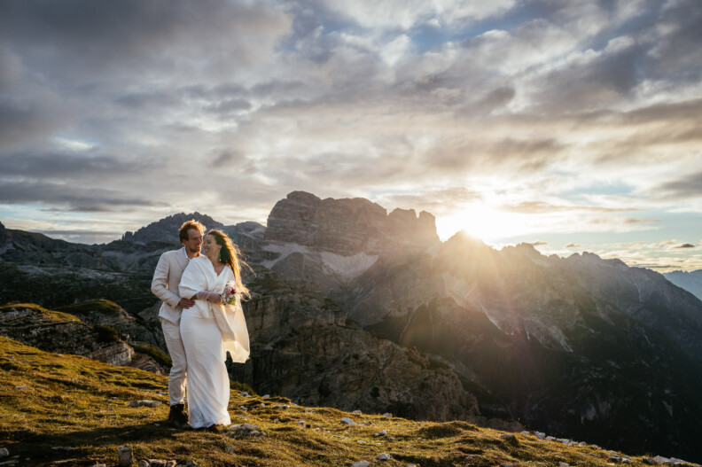 A couple in formal attire stands on a mountain ledge during sunset, with dramatic clouds and rugged peaks in the background.