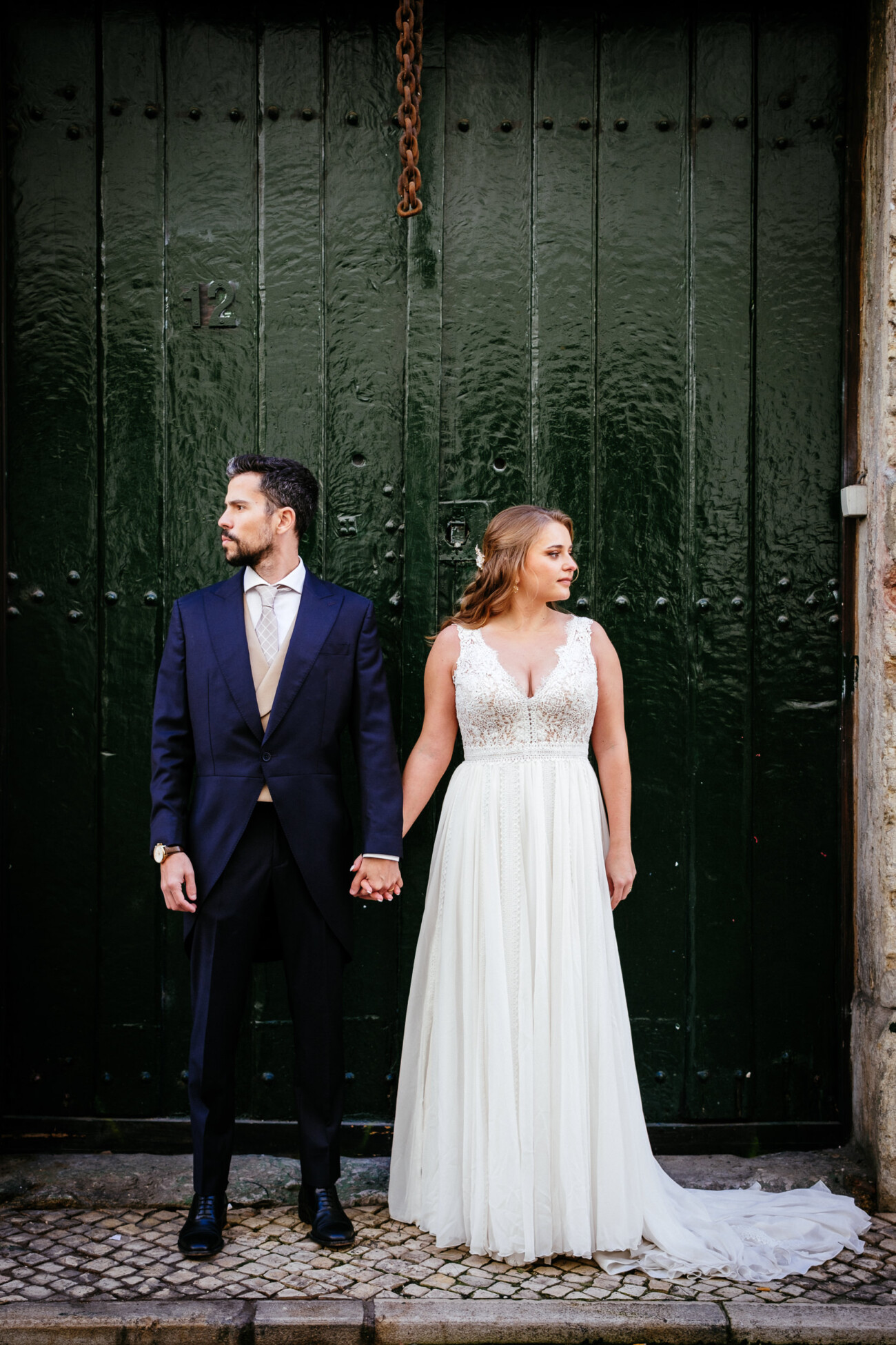 A couple in formal attire stand holding hands in front of a dark green, weathered door. The man is in a navy suit and the woman is in a white lace gown. They look in opposite directions.