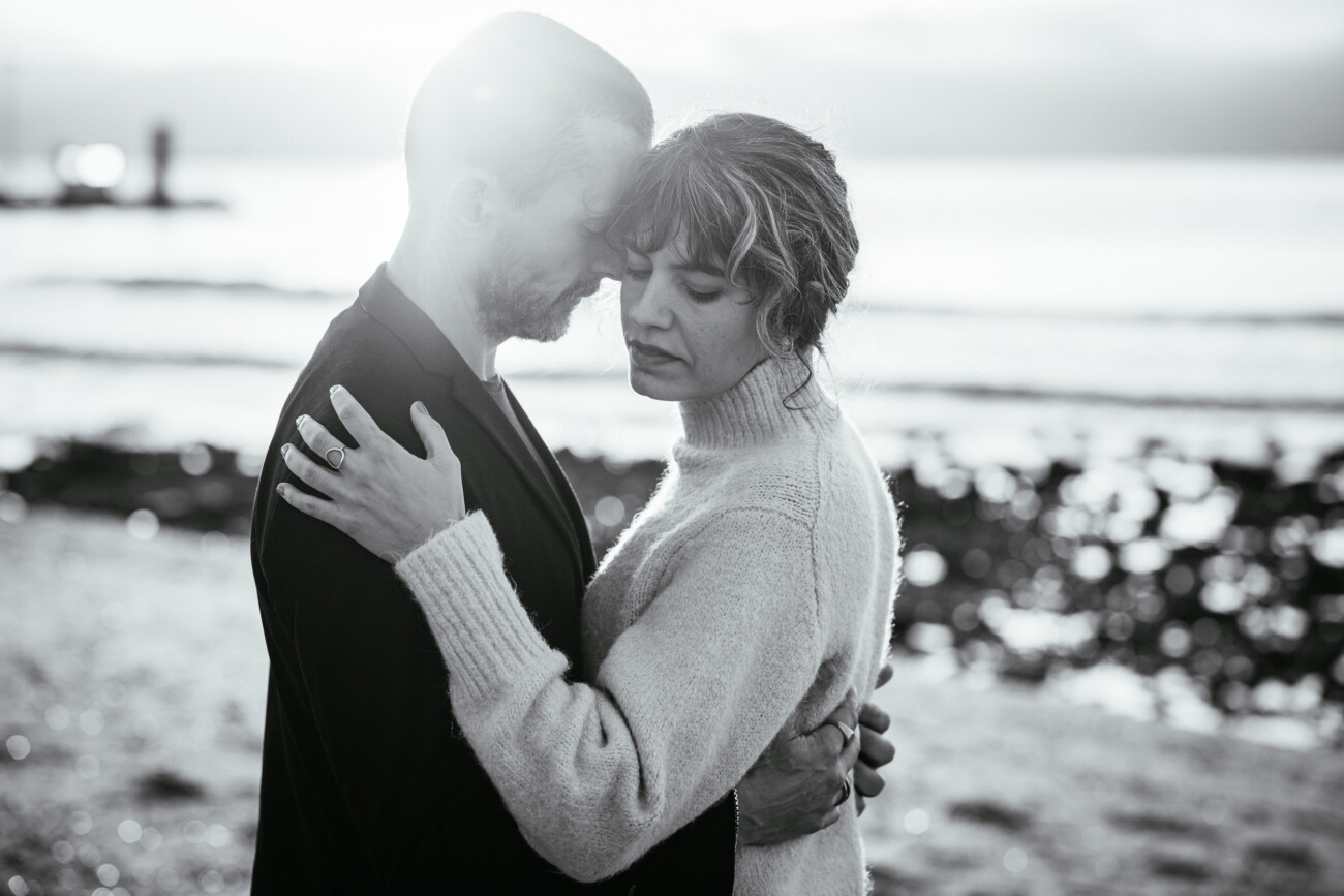 A couple embraces on a beach, their eyes closed, with the ocean in the background and sunlight illuminating the scene.