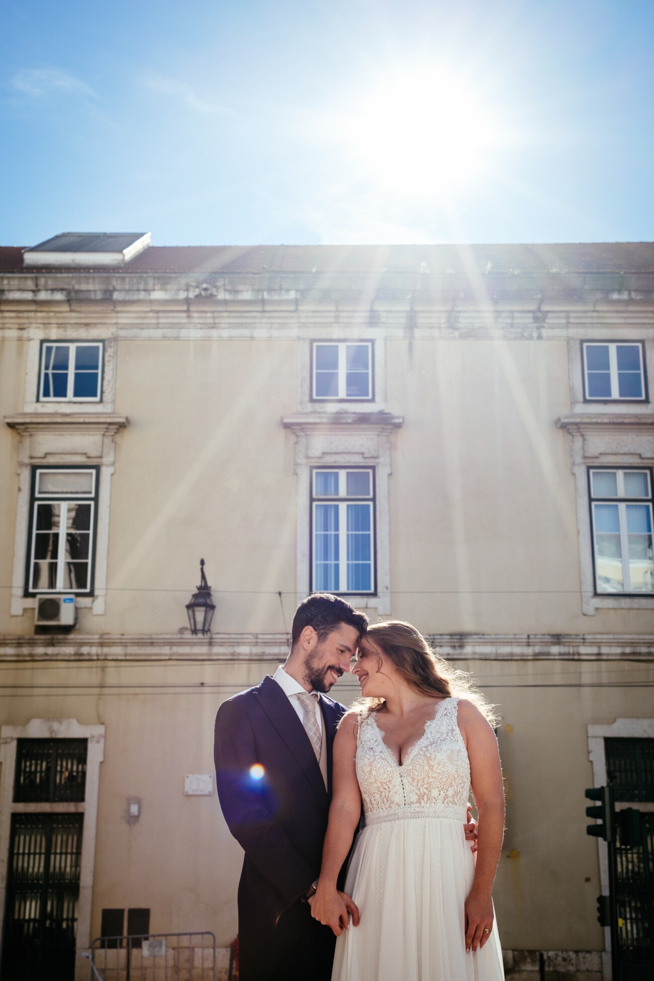 A couple in wedding attire stands in front of a beige building under a bright sunlit sky, sharing an affectionate moment.