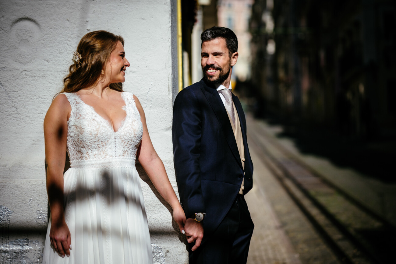 A bride and groom stand outdoors, holding hands and smiling at each other, next to a textured white wall with a street in the background.