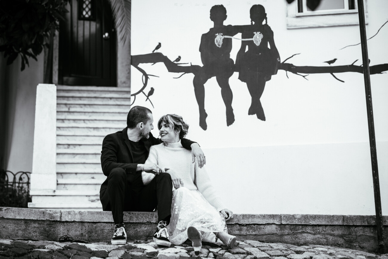 Couple sitting on cobblestone steps, smiling at each other. Black and white mural of two children with a string phone on the wall behind them.