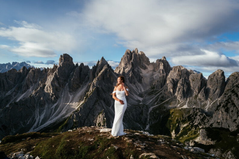 A woman in a white dress stands on a grassy hill with jagged mountains in the background under a cloudy sky.