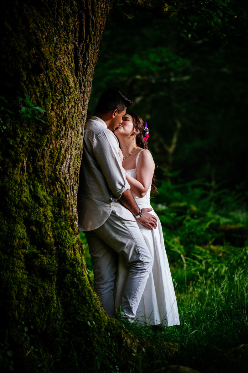 A couple in white attire stands by a large tree, embracing and sharing a kiss in a lush, green forest setting.