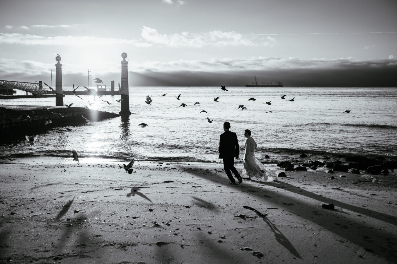 A couple walks along a beach at sunrise, surrounded by birds with a pier and distant ship visible in the background.