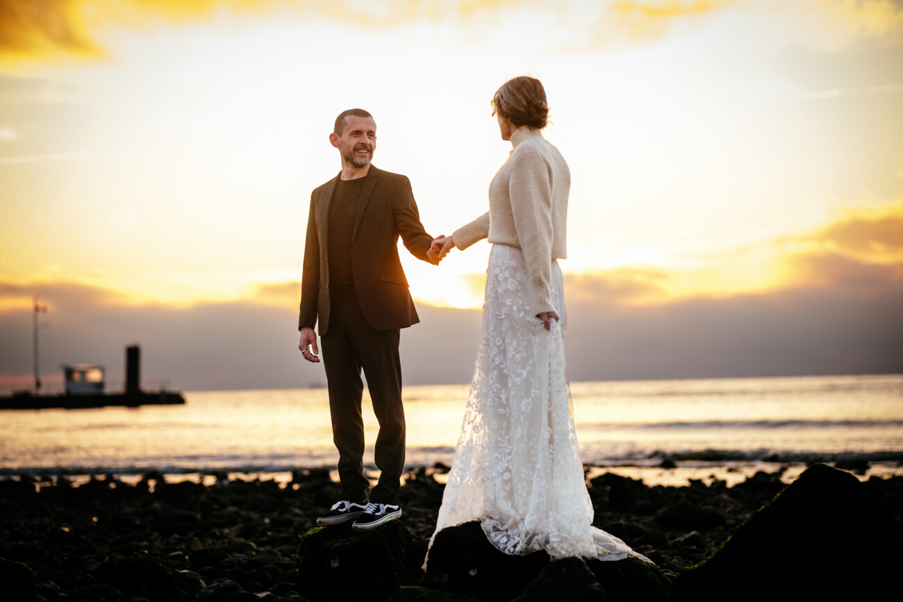 A couple stands on rocks by the sea at sunset, holding hands. The man wears a suit and the woman wears a long white dress with a sweater. A pier is visible in the background.