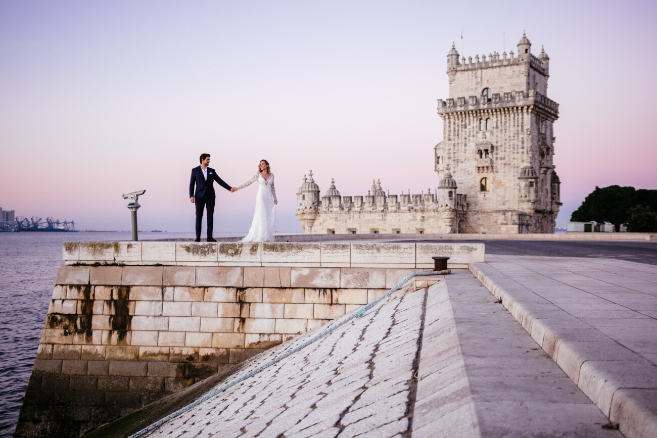 A couple in formal attire stands on the edge of a stone walkway with a historic tower and calm water in the background during sunset.