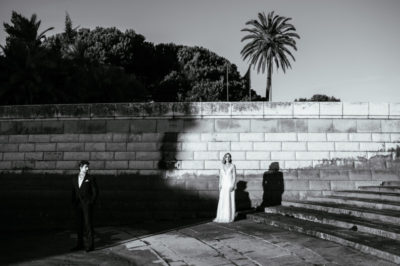 A bride and groom stand apart on stone steps with a palm tree in the background. The scene is in black and white.