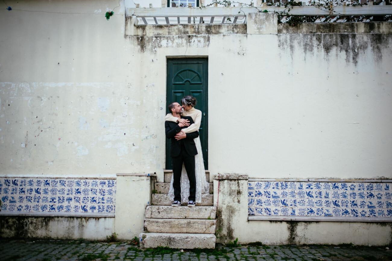 A couple stands embracing on steps in front of a weathered building with blue tile accents and a green door.