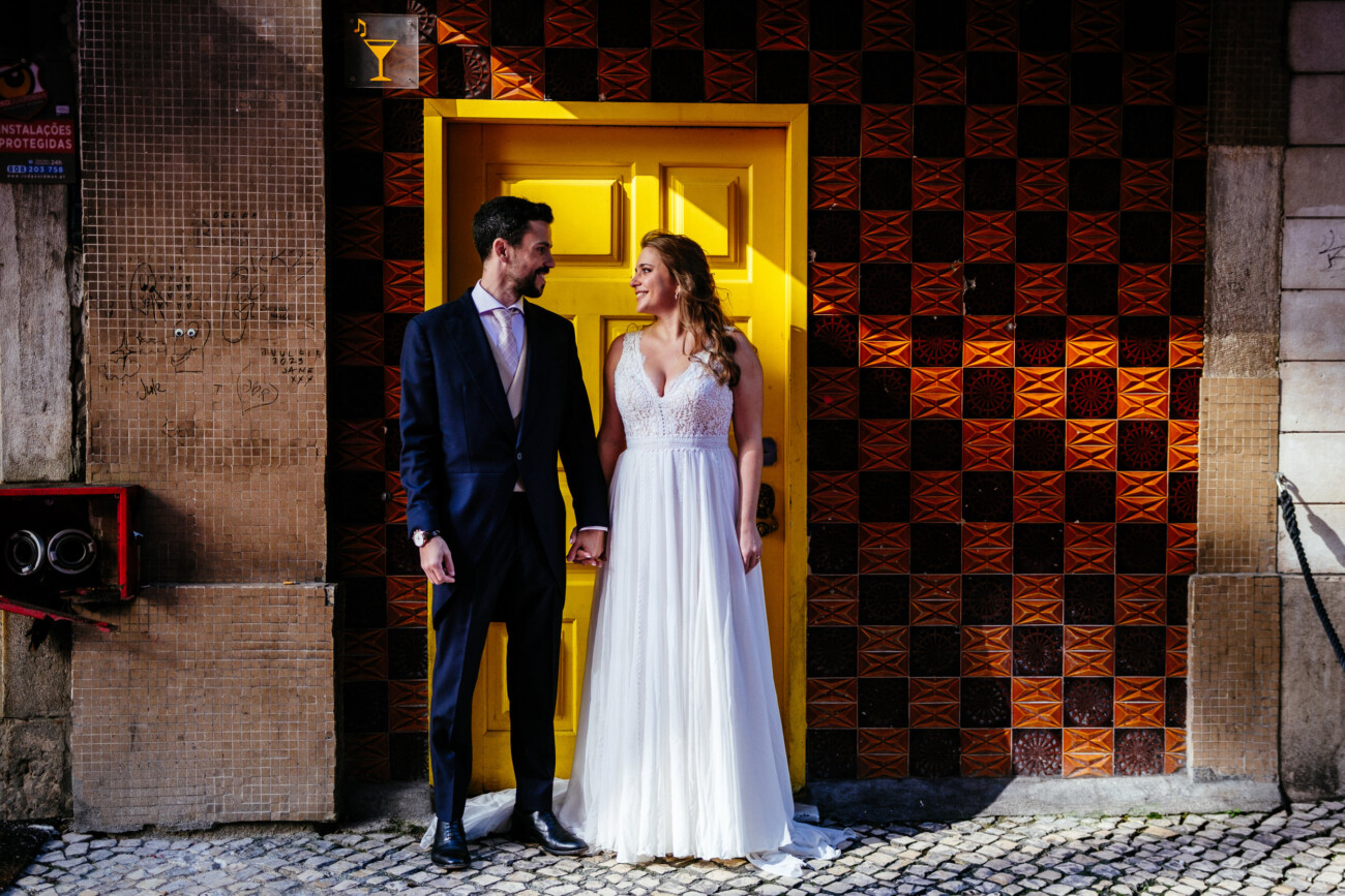 A couple in formal attire stands hand in hand in front of a bright yellow door, with patterned tiles on the wall behind them.