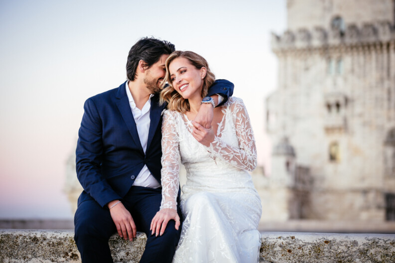 A couple in formal attire sits closely, smiling, with an ancient stone building in the background.
