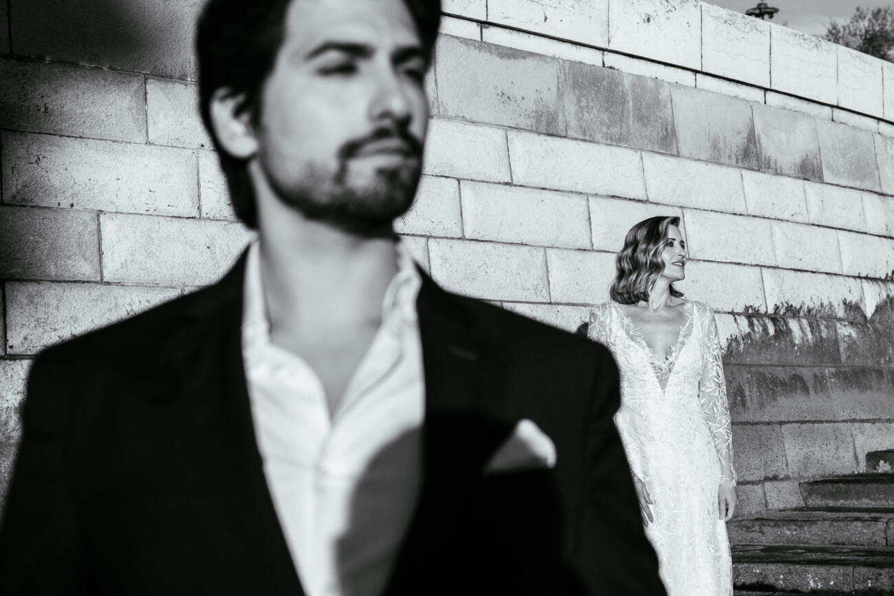 A man in focus stands in the foreground, wearing a suit, while a woman in a white dress stands in the background against a stone wall. The image is in black and white.