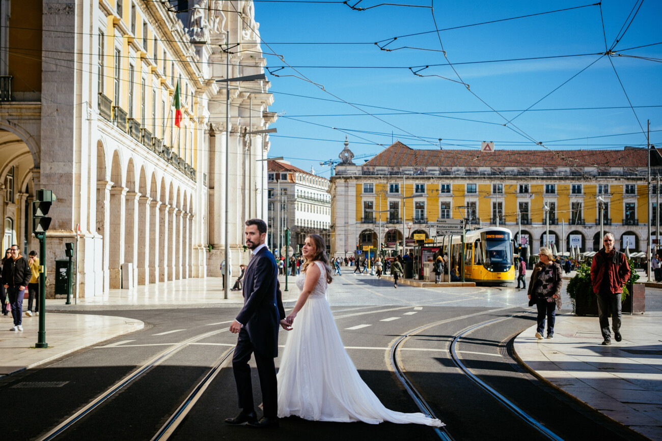 A couple in wedding attire holds hands while crossing a tram-lined street in a sunny, historic European plaza.
