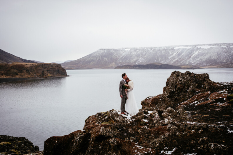 A couple in wedding attire stands on a rocky landscape by a lake, surrounded by mountains under an overcast sky.