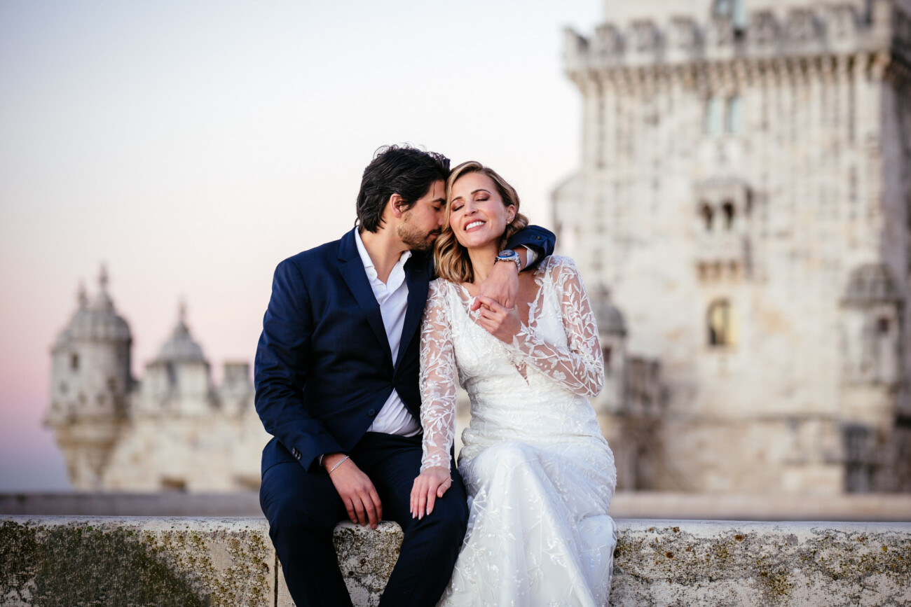A couple in formal attire sits on a stone ledge, smiling. The man is in a dark suit, and the woman wears a white dress. An ancient stone building stands in the background.