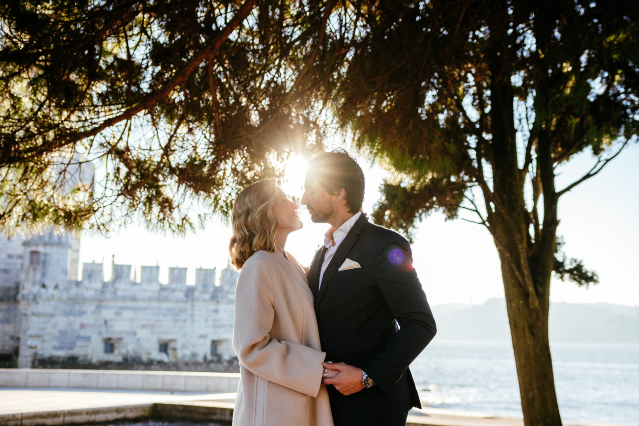 A couple in formal attire stands near a large tree, with sunlight filtering through the branches, and a stone building and water in the background.