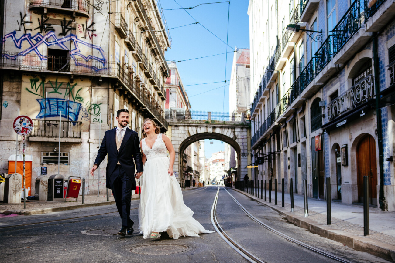 A couple in wedding attire walks joyfully down an urban street, surrounded by buildings with graffiti and balconies.