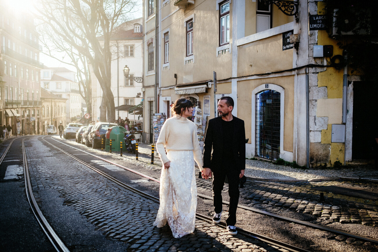 A couple holding hands walks along a cobblestone street lined with buildings and parked cars.