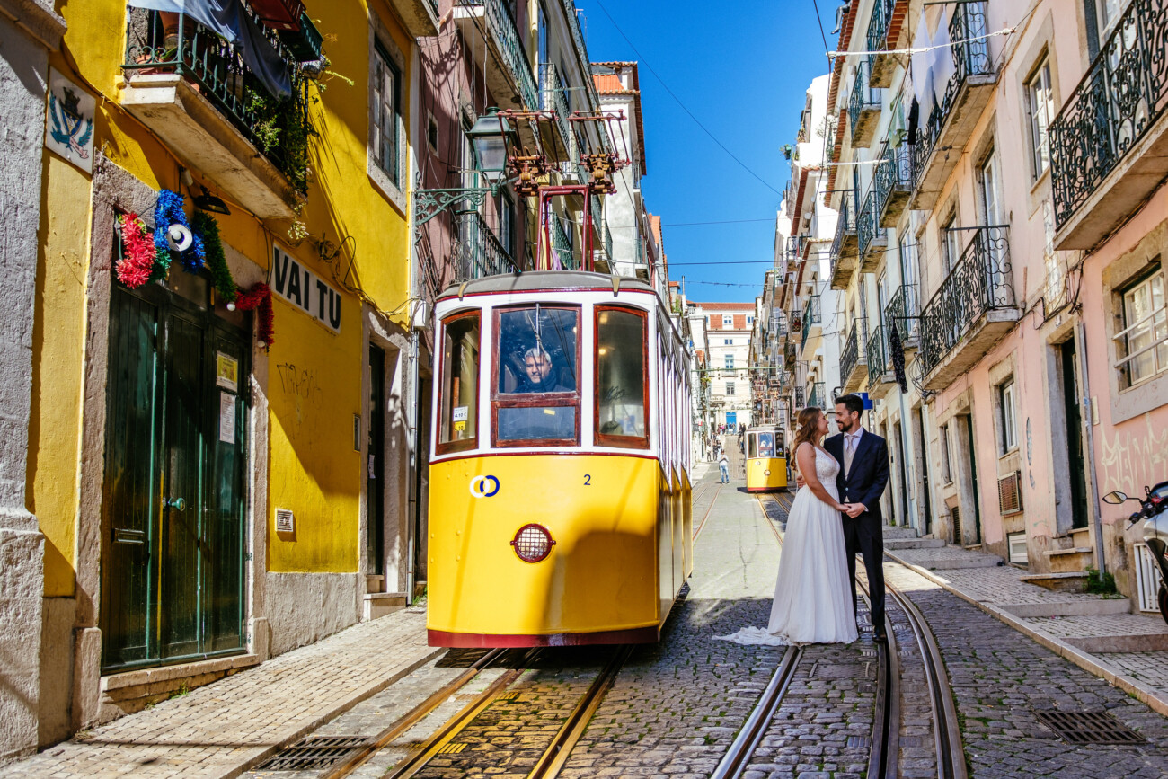 A couple in wedding attire poses near a yellow tram on a steep, narrow street lined with colorful buildings under a clear blue sky.