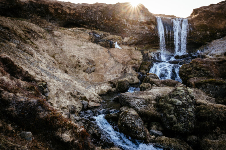 A couple stands on rocky terrain near a waterfall, with sunlight peeking over the cliff.
